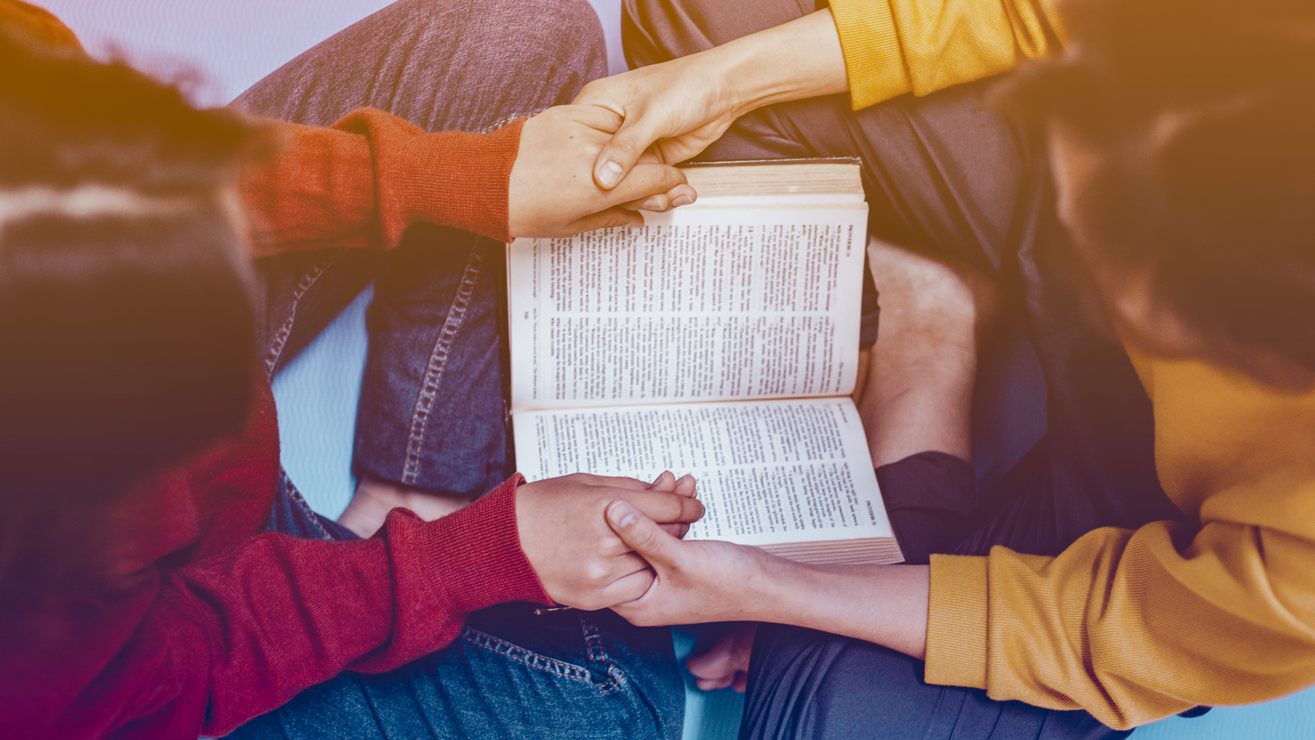 two young people hold hands across an open bible as if praying.
