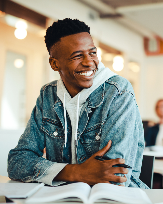 a young african american man sits in a classroom, smiling. This indicates he is undergoing some good leadership development.